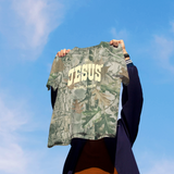 Person holding a camouflage shirt with 'JESUS' printed on it against a blue sky.