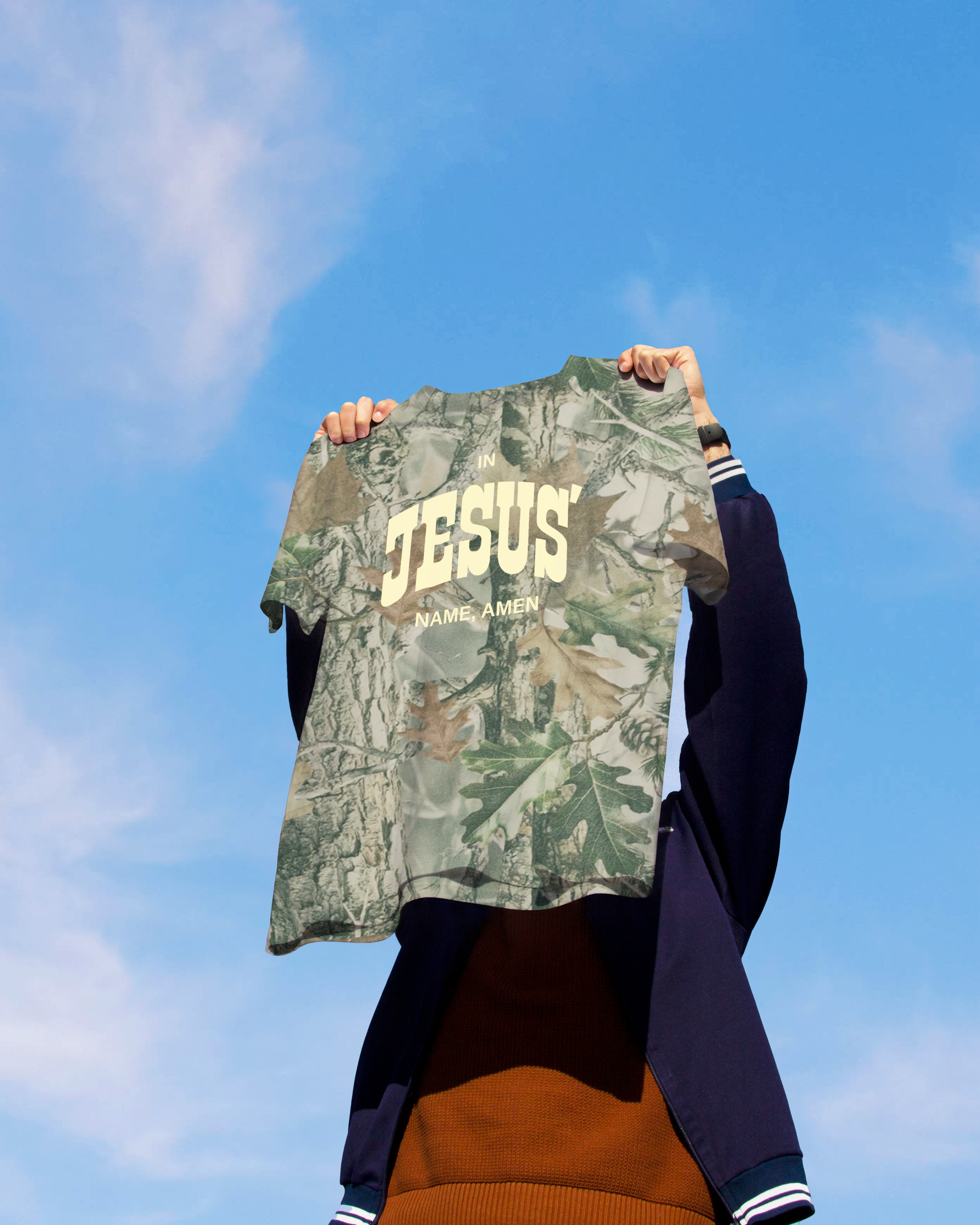 Person holding a camouflage shirt with 'JESUS' printed on it against a blue sky.
